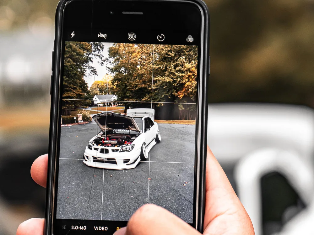 Kéz mobiltelefonnal fényképez egy fehér autót nyitott motorháztetővel, a kijelzőn az autó képe